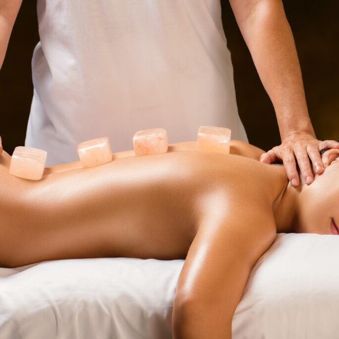 Close up of young woman having hot himalayan stone massage in spa.Natural salt stone bricks placed on spine with therapist in background.